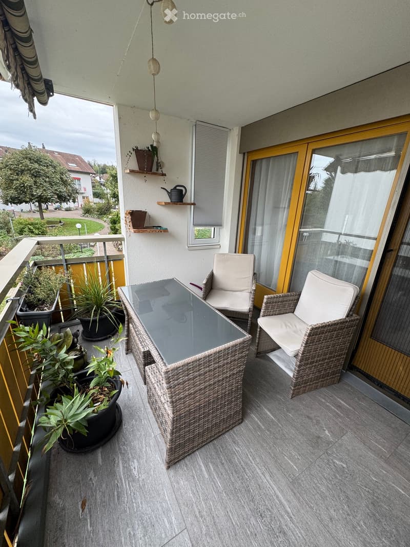 Balcony with wicker furniture, glass table, two chairs, and several potted plants. Features a view of a garden and a house in the background.