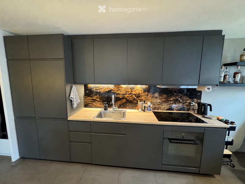 Modern kitchen with dark grey cabinets, wooden countertop, stainless steel sink, induction cooktop, and built-in oven. Features a decorative backsplash with a textured design.