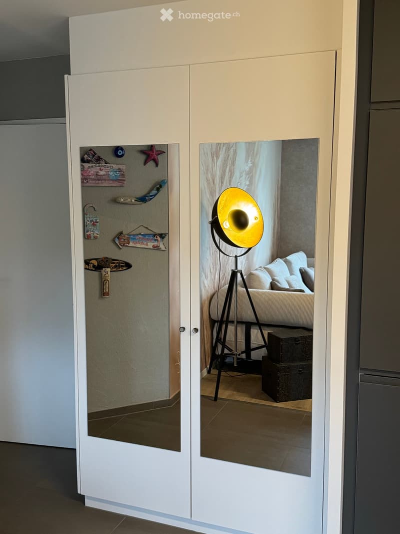 A modern hallway with white sliding doors, one leading to a decorated wall with various decorative items, and the other reflecting a cozy living space with a sofa, pillows, a floor lamp, and a wooden chest.