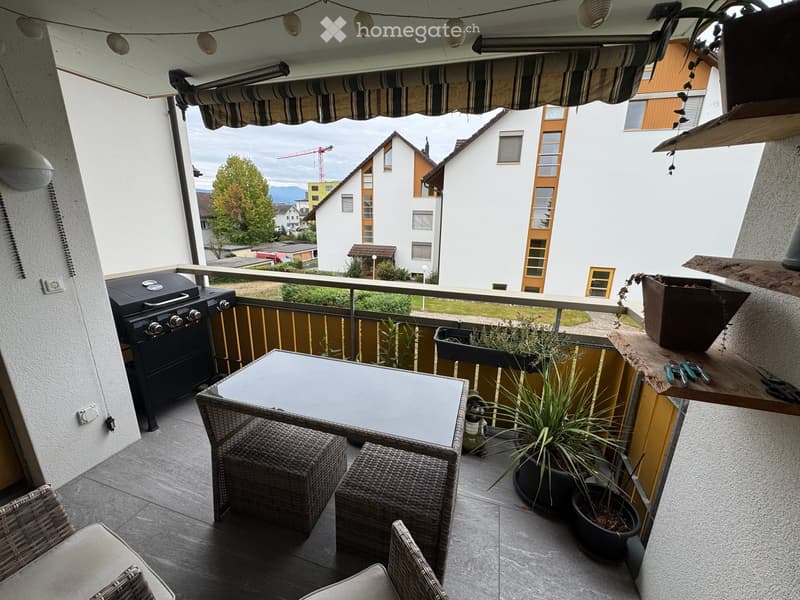 Balcony with a table, chairs, stools, grill, and potted plants, overlooking a residential area with houses and trees.