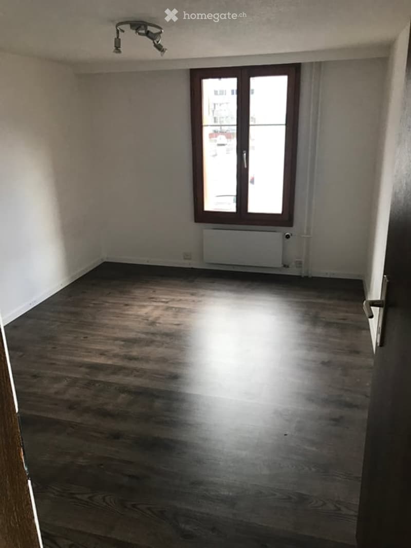 Empty room with wooden flooring, white walls, and a large window with brown frame. No furniture or equipment present.