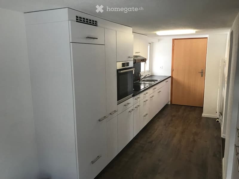 Modern kitchen with white cabinets, stainless steel oven, sink, and faucet. Dark wooden flooring and a wooden door visible.