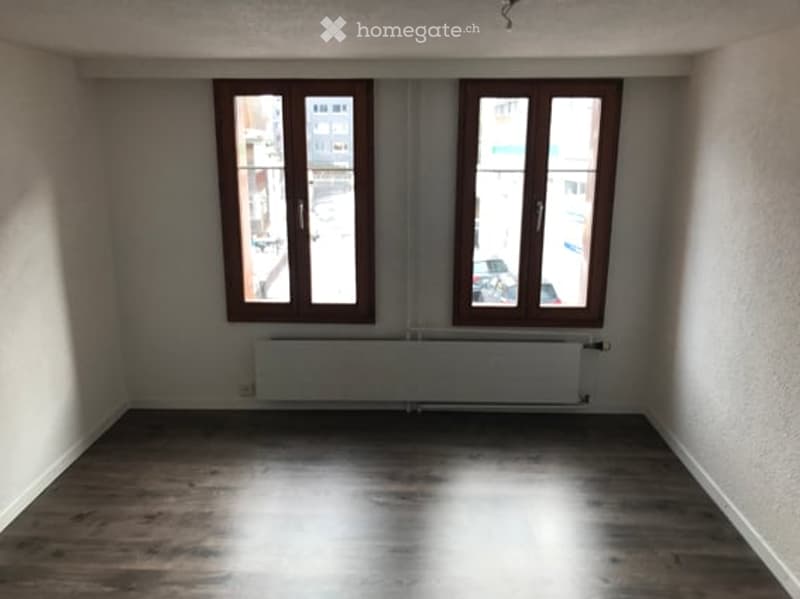 Empty room with wooden flooring, white walls, and two windows with brown frames. A radiator is visible below the windows.