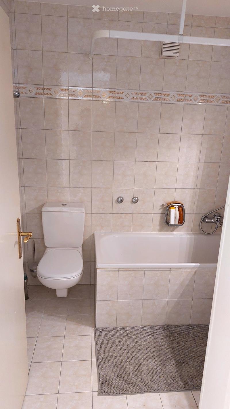 Bathroom with beige tiled walls, a white toilet, a bathtub with a safety rail, a small shelf with toiletries, and a grey bathmat on the floor.