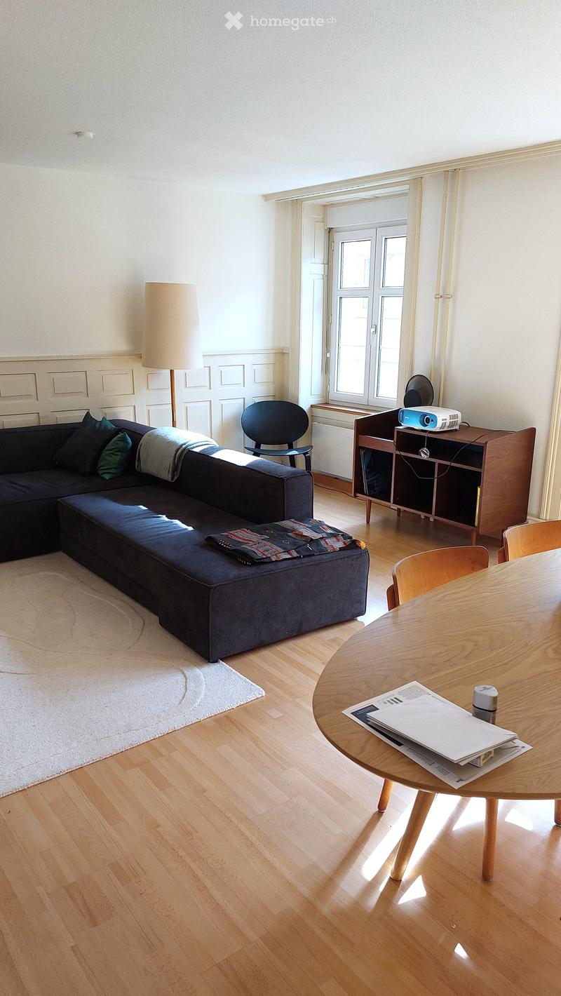 Bright living room with wooden flooring, a dark blue sectional sofa, a round wooden table, a floor lamp, and a projector on a wooden stand.