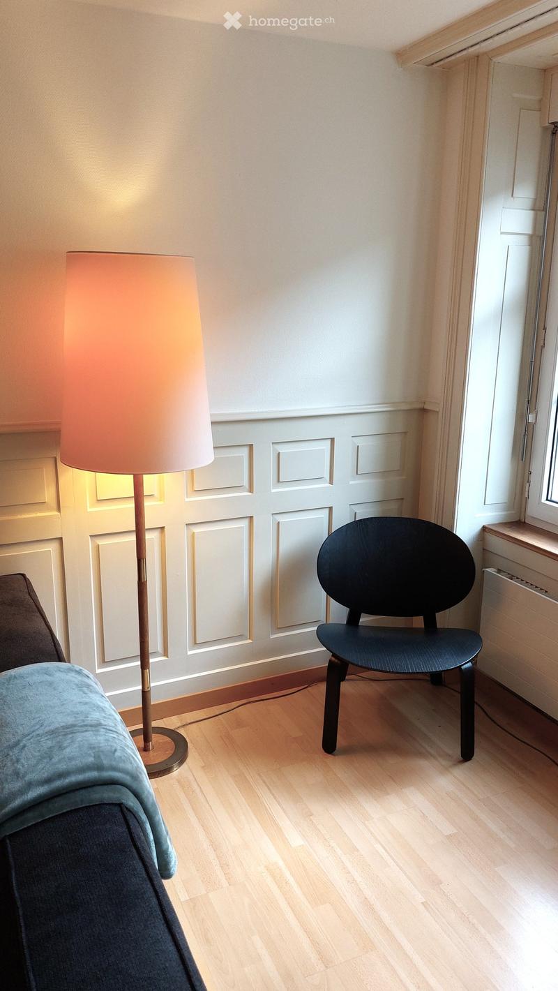 A small, modern room with light wooden flooring, a grey sofa, a black chair, and a tall floor lamp with a beige shade.
