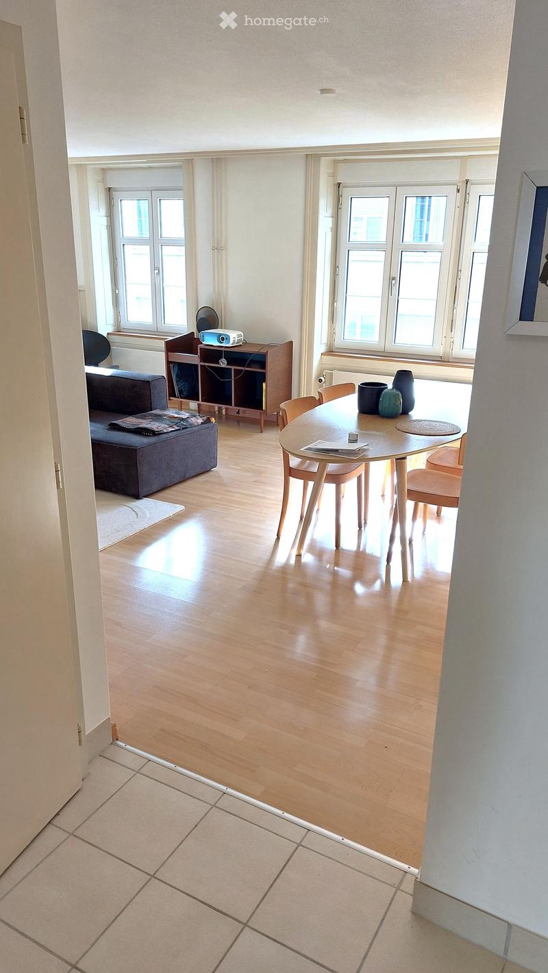 Bright living and dining area with wooden flooring, large windows, a sofa, a dining table with chairs, a shelf with a projector, and decorative vases.