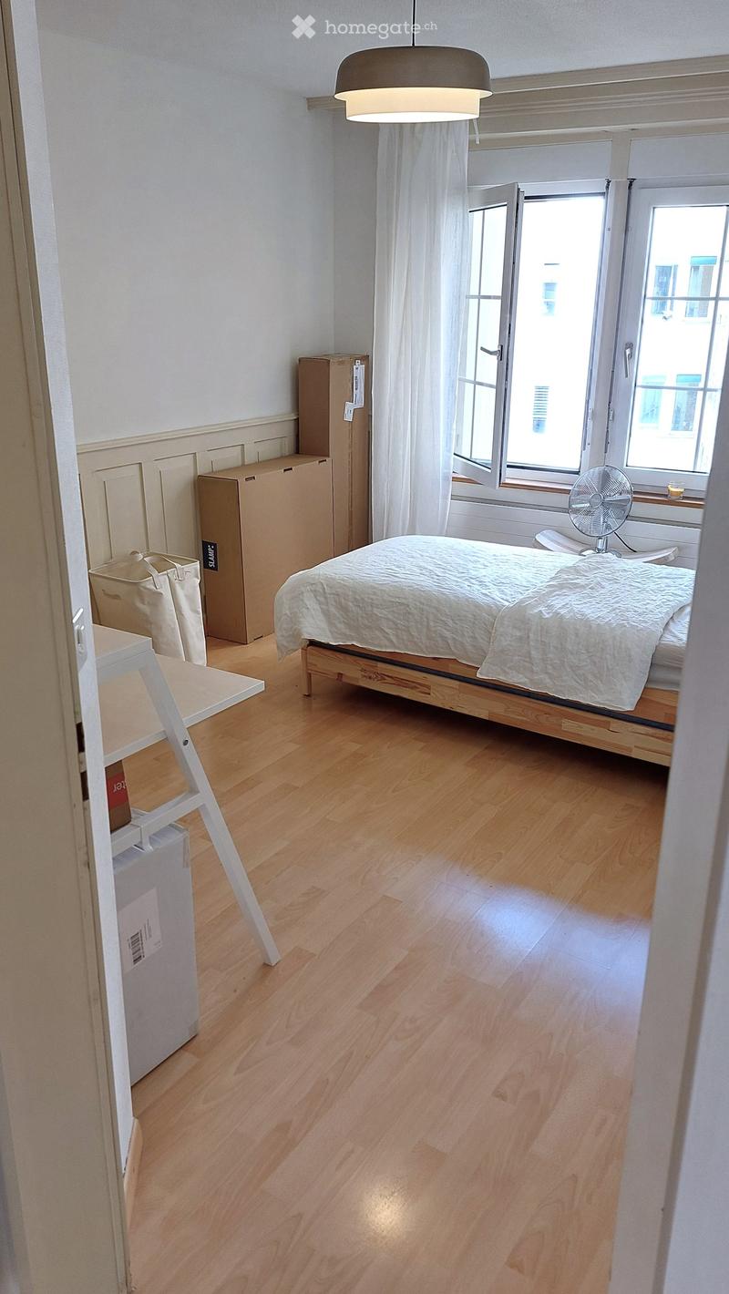 Bedroom with wooden floor, bed with white bedding, fan near window, and open window with white curtains.