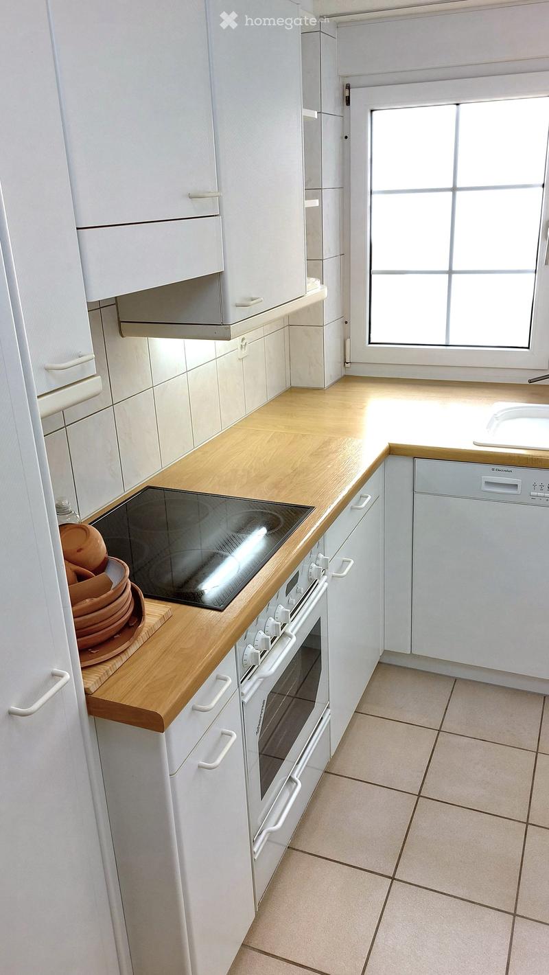 Modern kitchen with white cabinets, light wooden countertops, and built-in appliances including an oven, dishwasher, and induction stove. A window provides natural light.