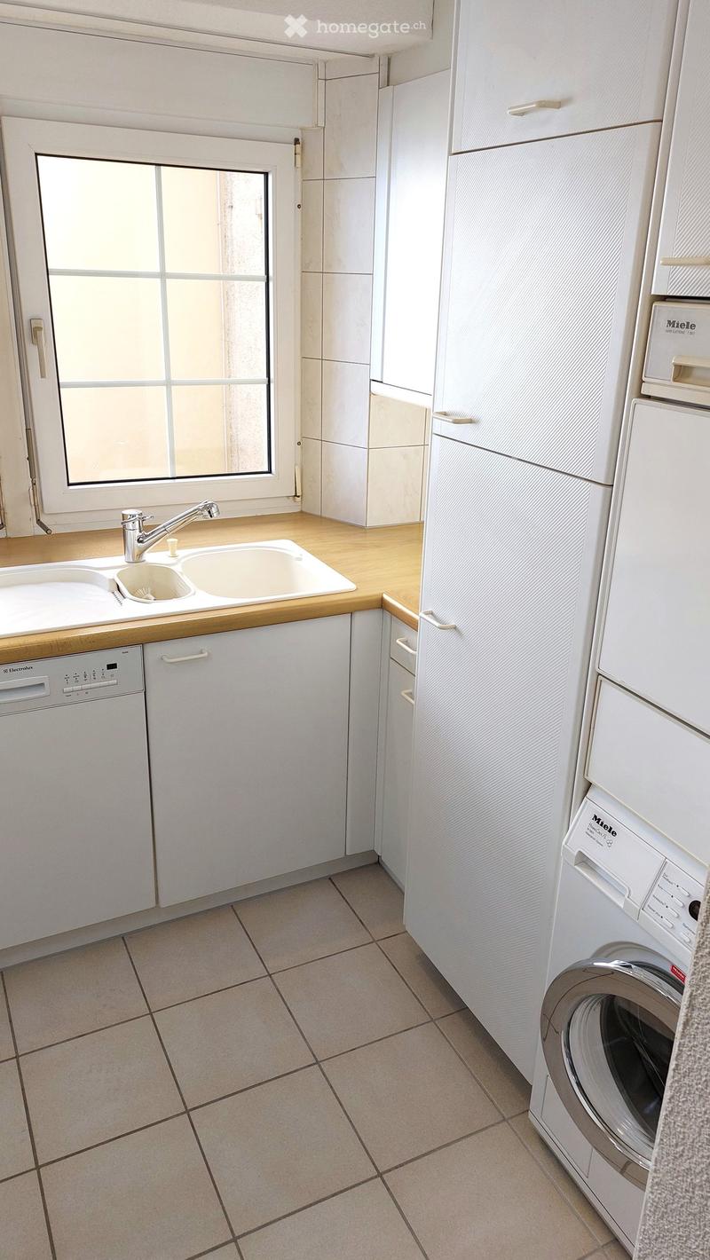 Modern kitchen with white cabinets, a double sink, a dishwasher, a refrigerator, and a washing machine. The floor is tiled, and there is a window with a grid pattern above the sink.