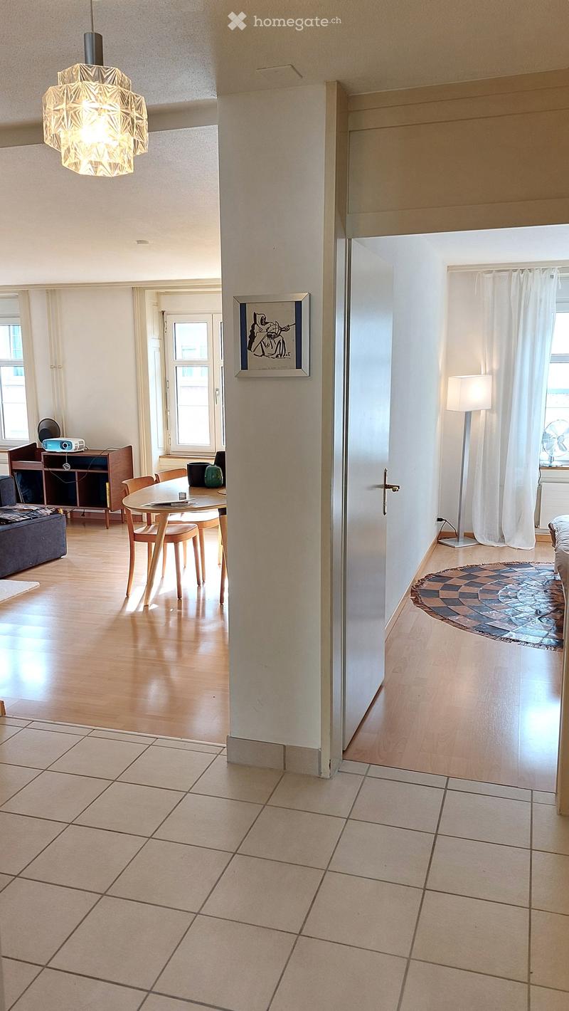 Open-plan living and dining area with wooden flooring, a sofa, a dining table with chairs, a lamp, a picture frame on the wall, and a fan near the window.
