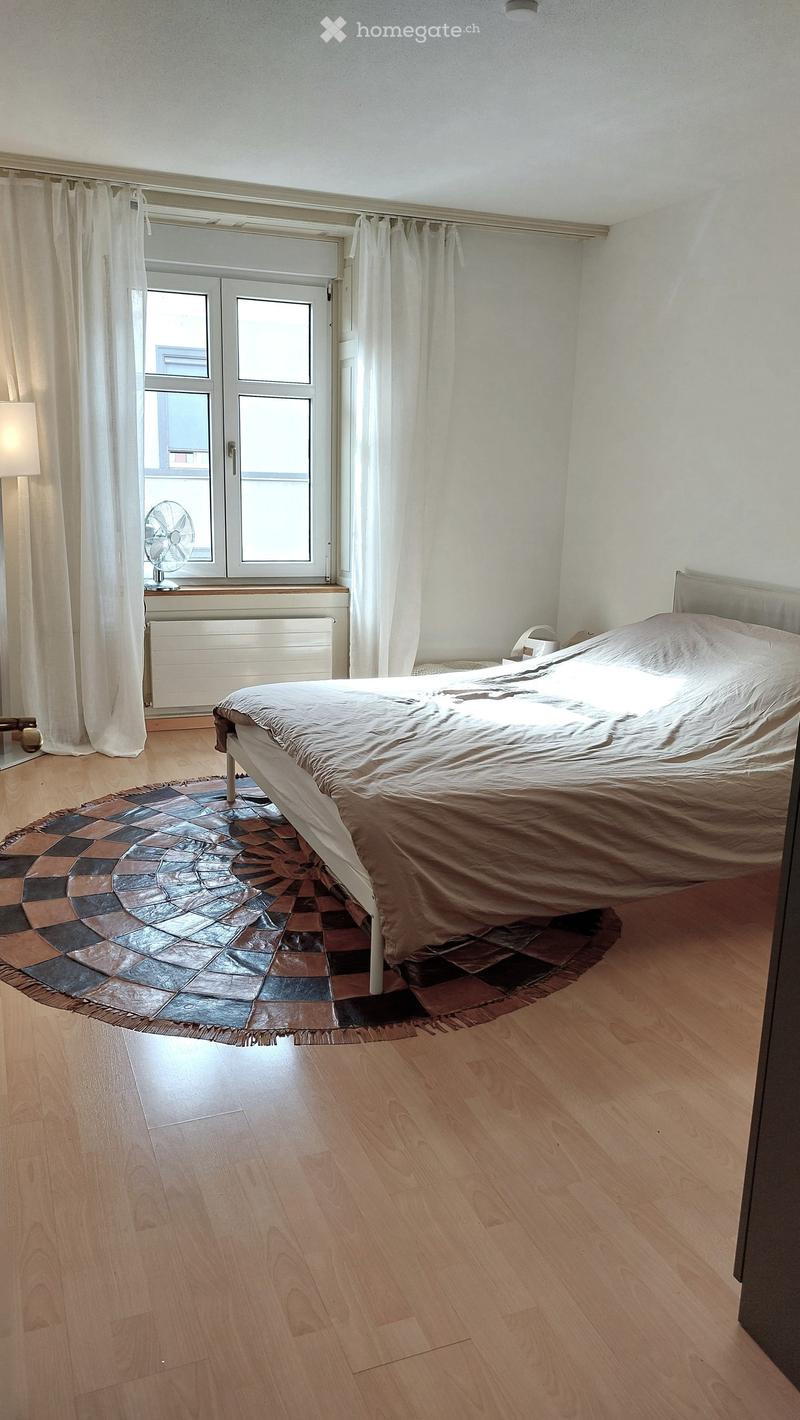 Bedroom with wooden flooring, a large bed with beige bedding, a round patterned rug, a fan on the windowsill, and a lamp near the bed.
