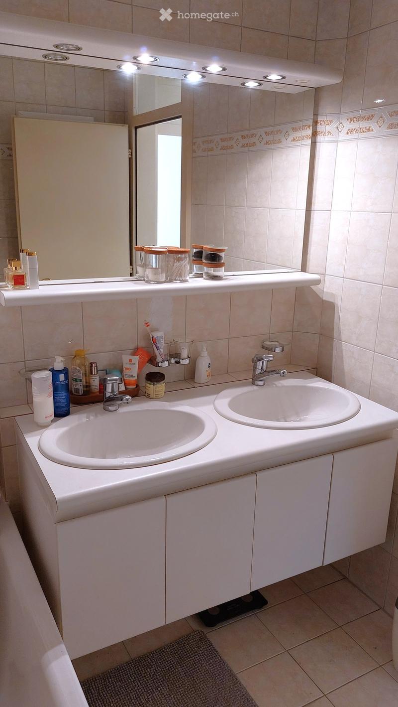 Modern bathroom with double sink, mirror with built-in lighting, various toiletries, and a small rug on the floor.
