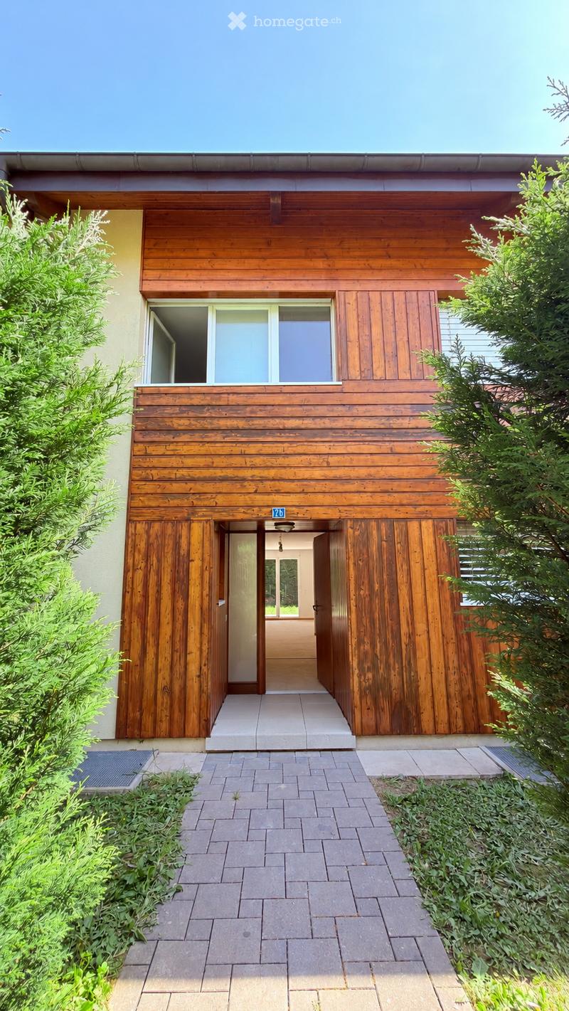 Modern two-story house with wooden exterior, large windows, and a covered entrance area. The house features a raised ground floor and is surrounded by greenery and a paved pathway.
