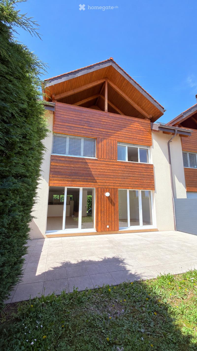 Modern two-story house with wooden exterior, large windows, and a raised ground floor. Features a balcony on the upper level and a well-maintained garden in the foreground.