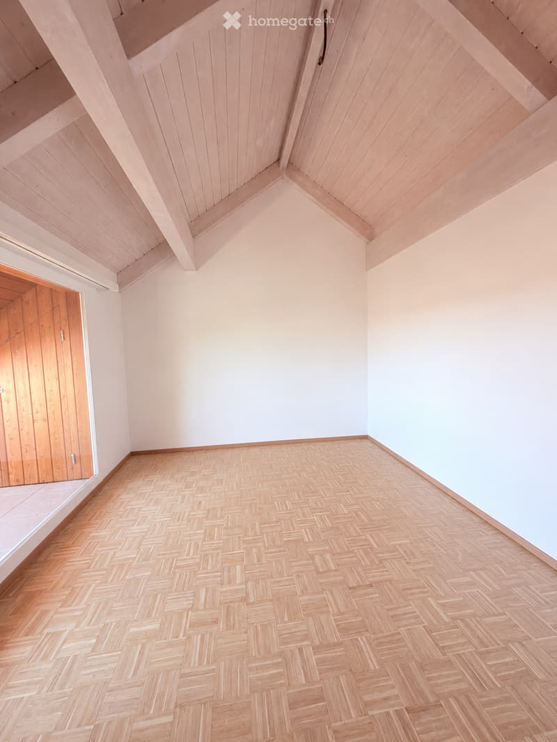Empty room with wooden herringbone flooring, light-colored walls, and a slanted wooden ceiling. A wooden door is visible on the left side.