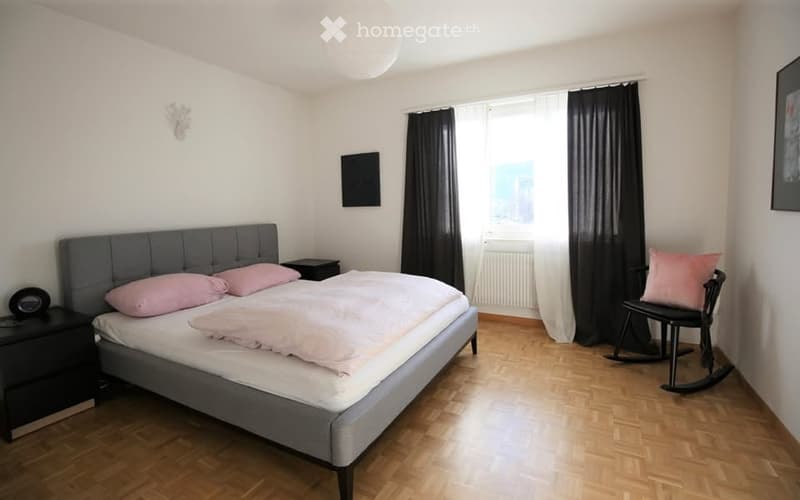 Modern bedroom with a grey upholstered bed, pink pillows, a nightstand, and a rocking chair. The room features wooden flooring, large windows with black curtains, and a decorative wall piece.