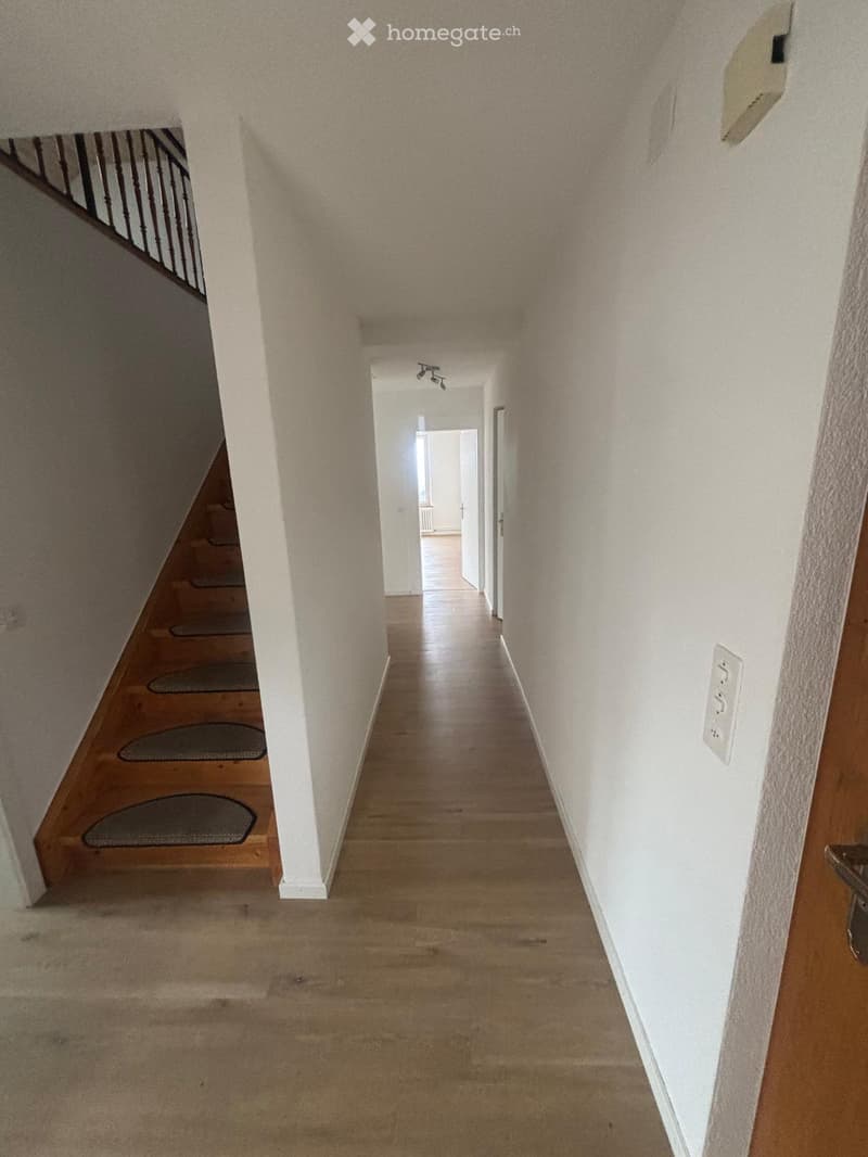 A long, well-lit hallway with wooden flooring, white walls, and a staircase on the left side. The hallway features a series of small, dark-colored rugs placed on the wooden stairs and a light fixture on the ceiling.