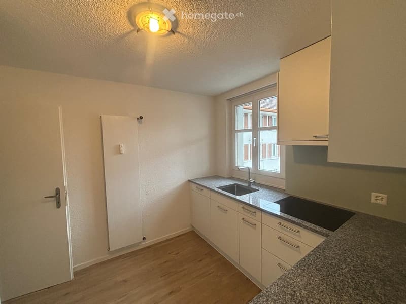 Modern kitchen with white cabinets, granite countertops, sink, and stove. Wooden flooring and large window provide natural light.