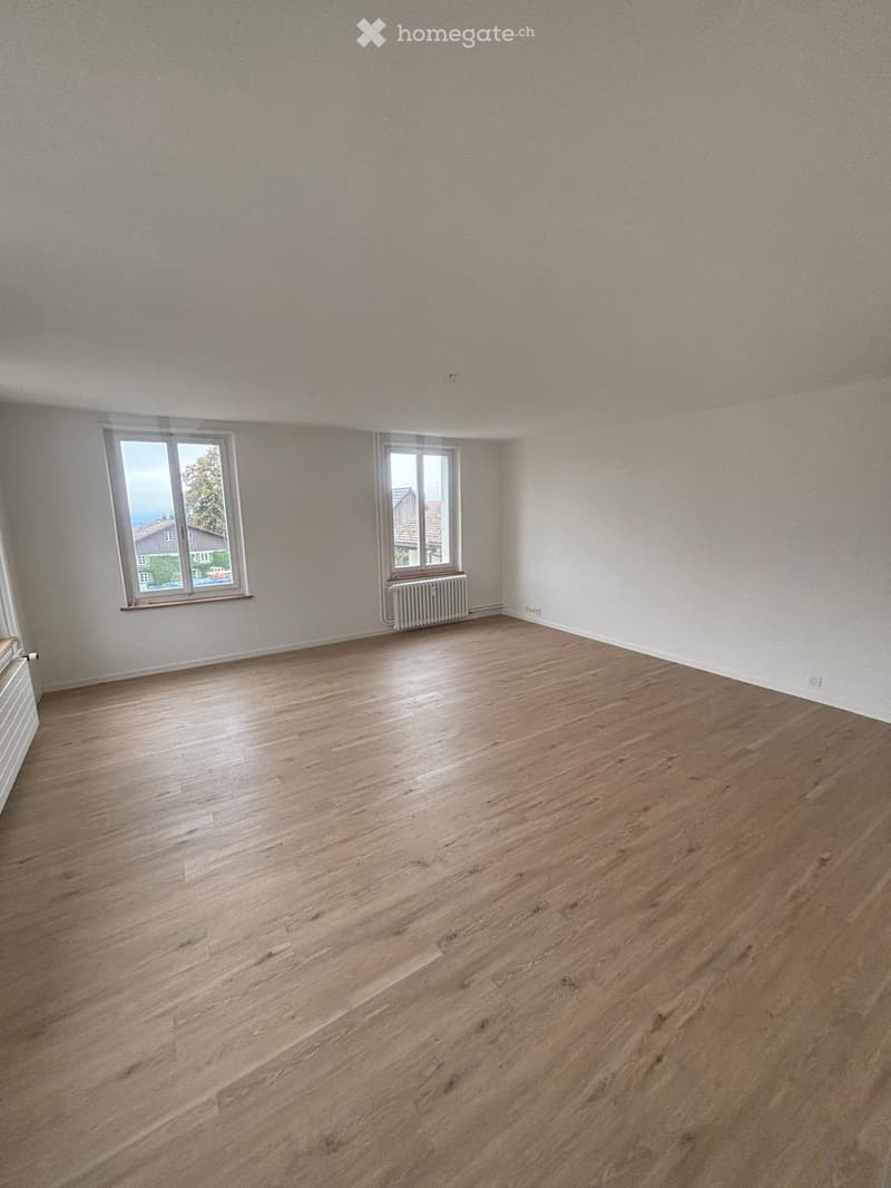 Empty room with wooden flooring, white walls, and two windows offering a view of the outside. Radiators are visible under the windows.