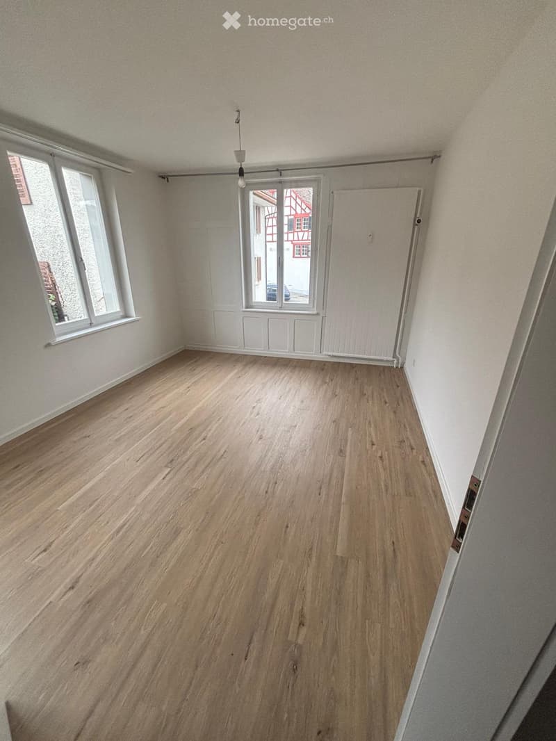 Empty room with wooden flooring, white walls, large windows, and a sliding glass door leading to a view of a traditional building.