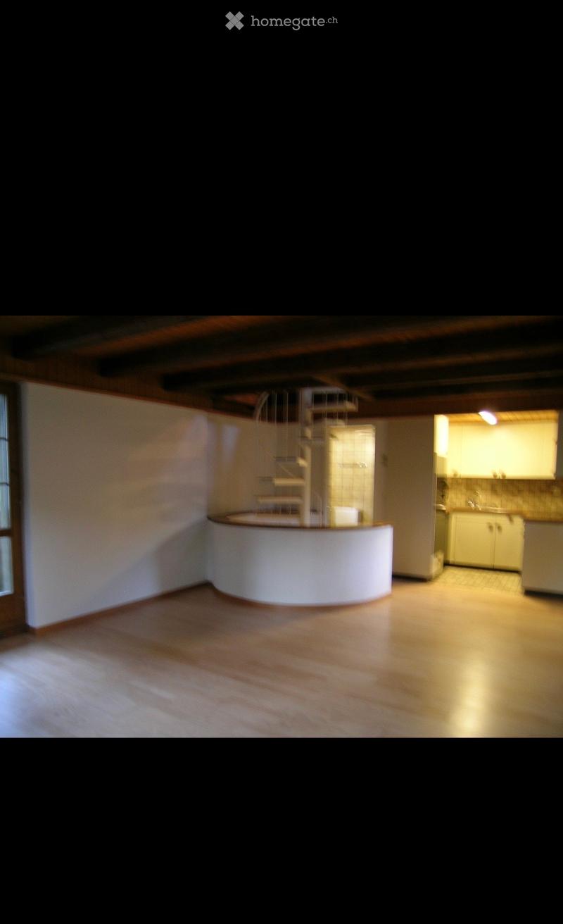 Empty room with wooden flooring, white walls, and a small spiral staircase leading to an upper level. The room is brightly lit with natural light coming from a window on the left side.