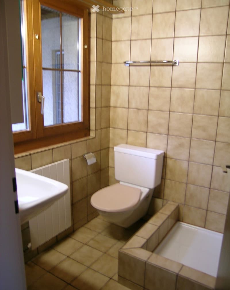 Bathroom with beige tiled walls and floor, toilet, sink, bathtub, and a window with wooden frame.