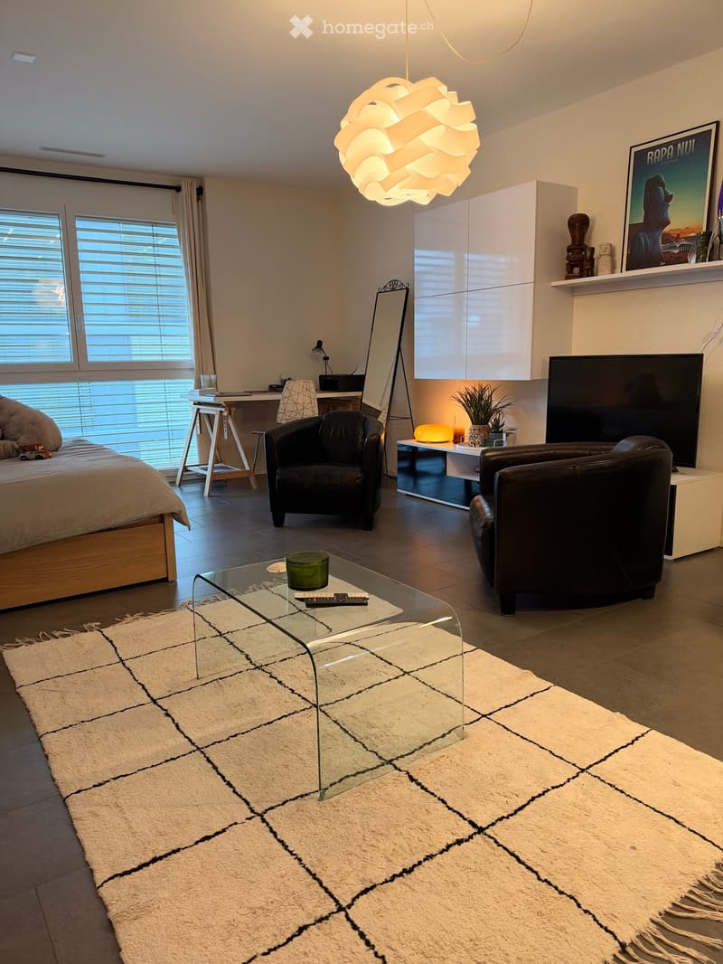 Modern living room with a large window, a white rug, a glass coffee table, two black leather chairs, a bed, a mirror, a small table with a lamp, and a TV screen.