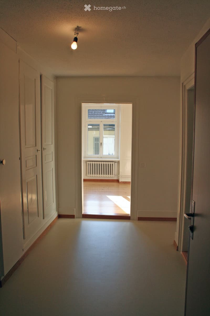 Empty hallway with light-colored walls, wooden flooring, and a single ceiling light fixture. A door on the left leads to another room, and a window at the end provides natural light.