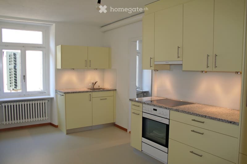 Bright, modern kitchen with light yellow cabinets, granite countertops, a sink, and an oven. Features large windows and a radiator under one window.
