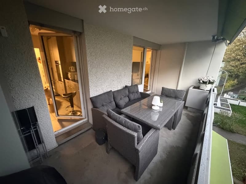 Balcony with outdoor furniture including a sofa, table, and chairs, with views of the interior kitchen and living area through large glass doors.