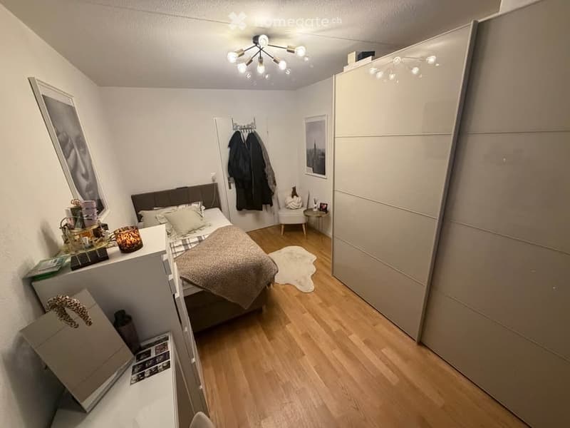 Cozy bedroom with wooden flooring, a double bed with pillows and a blanket, a nightstand with various items, a white chair, a large wardrobe, and a coat rack with jackets.