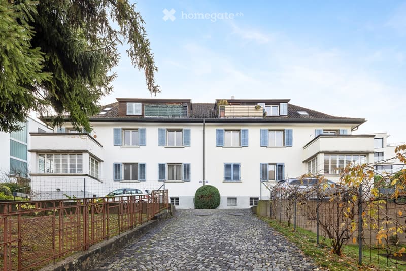 A two-story residential house with white walls, blue shutters, and a tiled roof. The house features multiple windows, a small balcony on the roof, and a gated entrance leading to a cobblestone pathway.