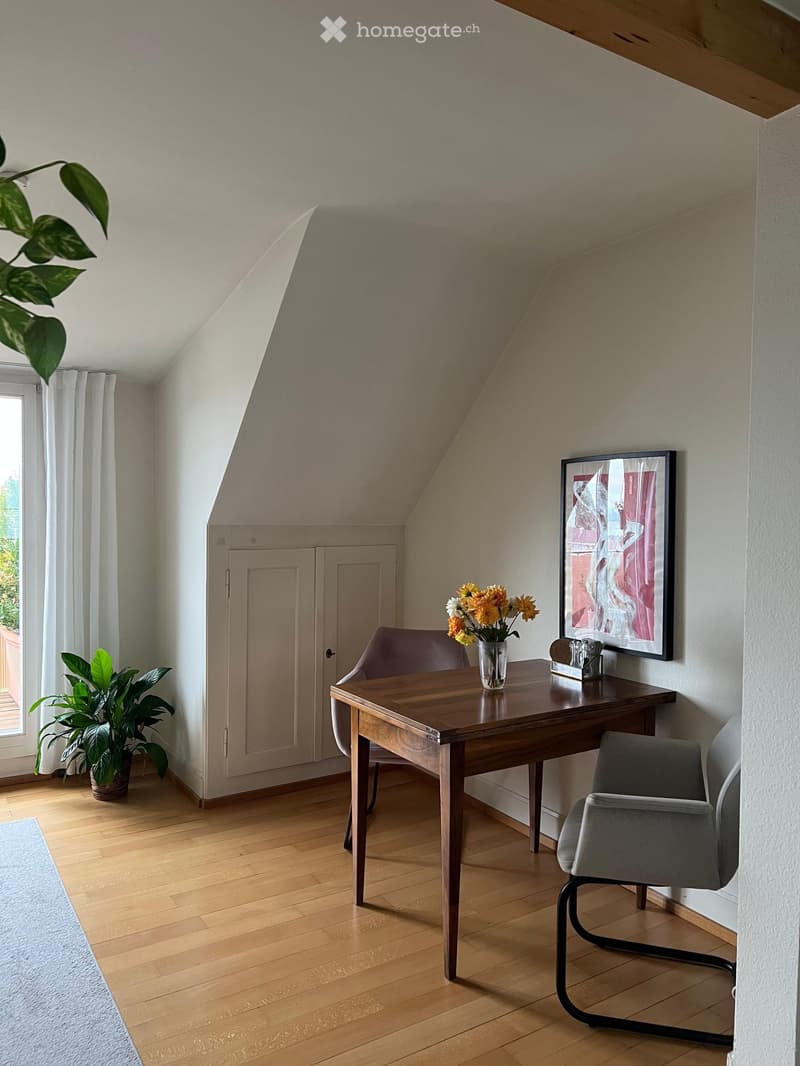 A cozy attic room with wooden flooring, a slanted ceiling, a wooden table with a vase of flowers, a grey chair, a plant, and a picture frame on the wall.