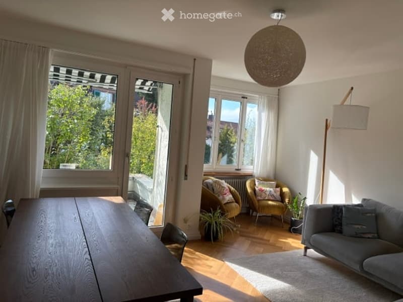 Bright living room with large windows, wooden flooring, grey sofa, dining table, armchairs, and a floor lamp.