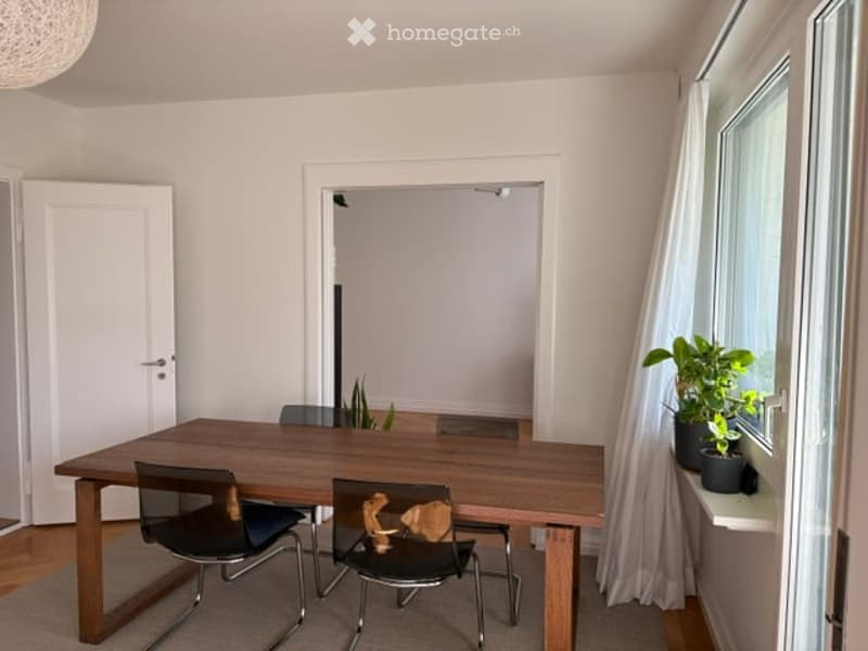 Bright dining area with a wooden dining table, four chairs, and a potted plant on the windowsill. The room has large windows with white curtains and a wooden floor.