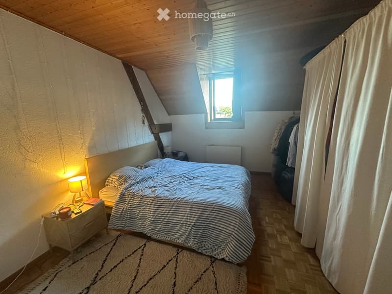 Cozy attic bedroom with wooden slanted ceilings, a large window, a bed with striped bedding, a nightstand with a lamp, and a wardrobe with hanging clothes.