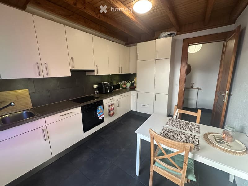 Modern kitchen with white cabinets, dark tiled floor, and a dining area with a white table and wooden chairs. The kitchen features a sink, oven, and refrigerator.