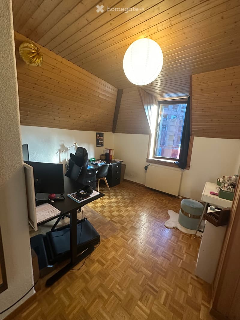 A cozy attic room with wooden slanted ceilings, a large window, a desk with a chair, a screen, and a small table with a stool.