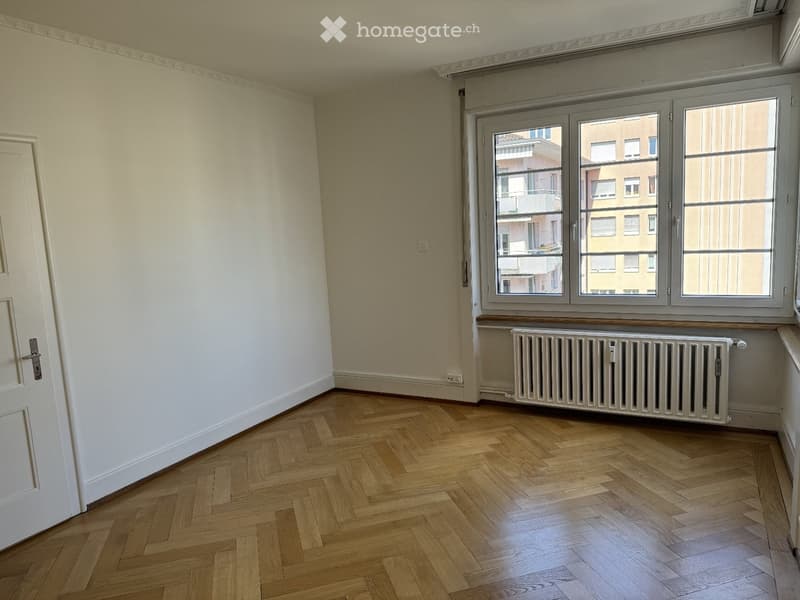 Empty room with wooden herringbone flooring, white walls, large window with radiator below, and a white door.