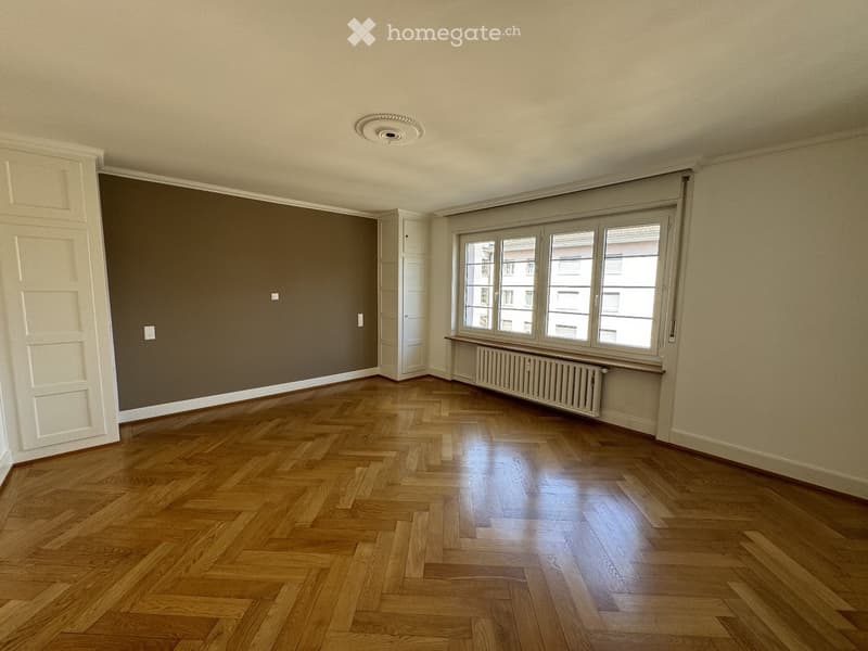 Empty room with wooden herringbone flooring, large window with radiator below, and neutral-colored walls.