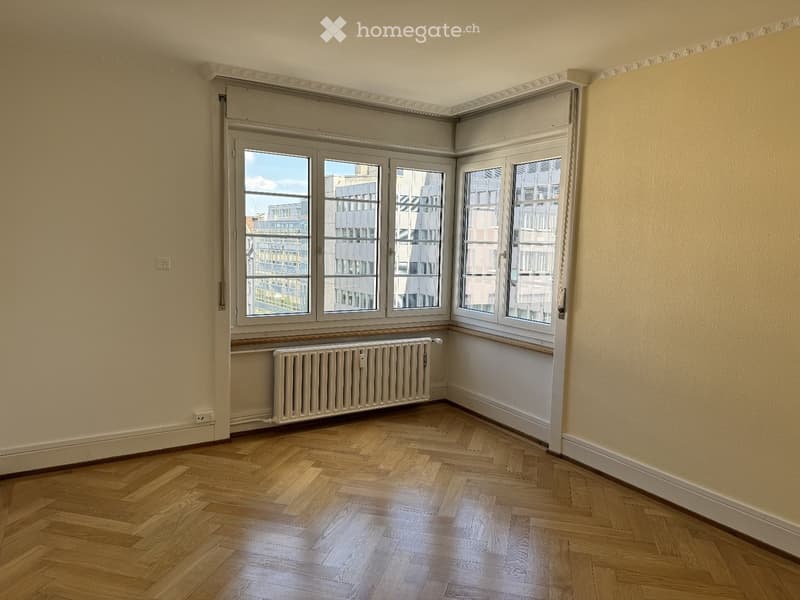 Empty room with wooden herringbone flooring, large windows with white frames, and a radiator below the windows.