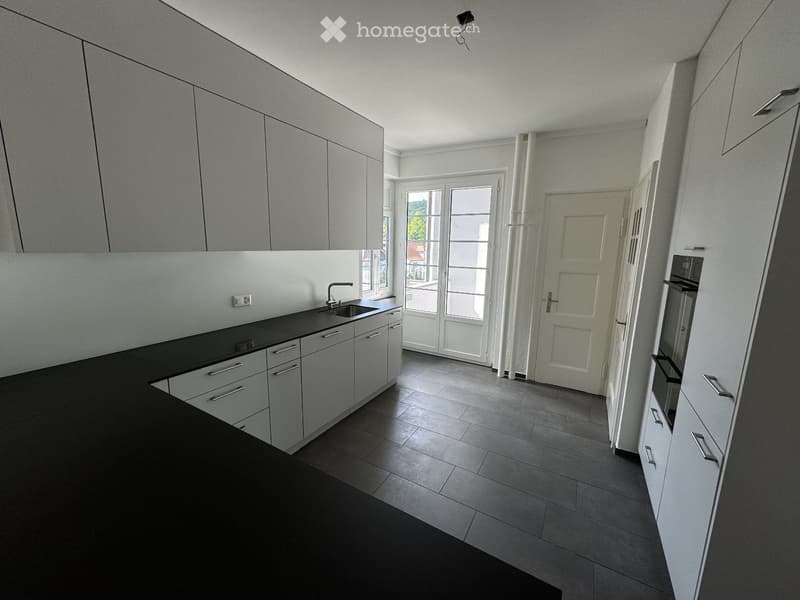 Modern, unfurnished kitchen with sleek white cabinets, black countertops, and a sink. The space features large windows and a door leading outside, with a clean and minimalist design.