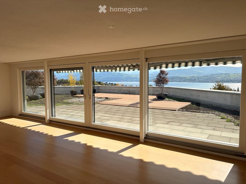 Bright, unfurnished room with large sliding glass doors opening to a wooden deck and scenic lake view.