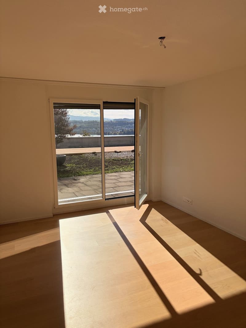 Empty room with wooden flooring, large sliding glass door leading to a balcony with a scenic view, and natural light streaming in.