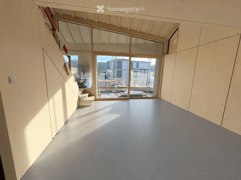 Empty room with light wooden walls, grey floor, large windows, and a staircase leading upwards.