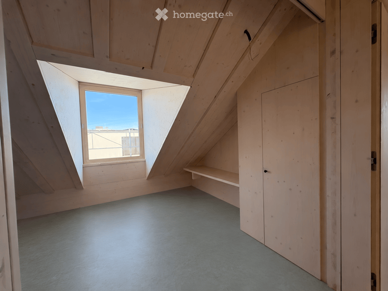 Empty attic room with wooden walls, slanted ceiling, and a single window offering a view of the sky and neighboring building.