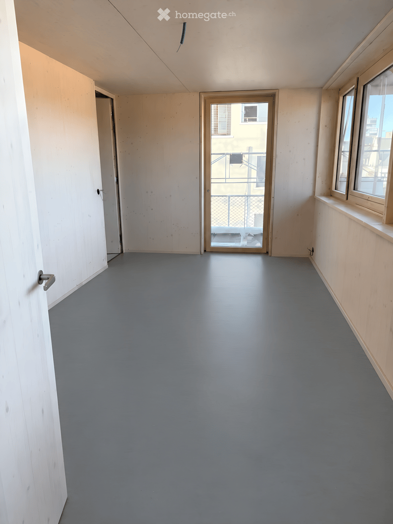 Empty room with grey flooring, light wooden walls, and a door leading to a balcony area.