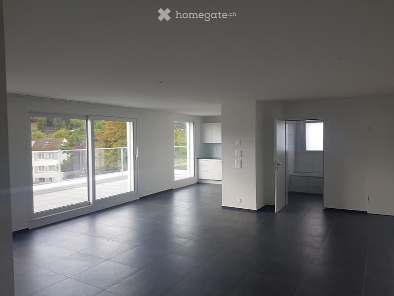 Empty room with dark tiled floor, large sliding glass doors leading to a balcony, and a small kitchen area visible in the background.