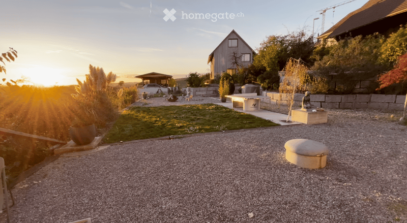 Well-maintained garden with gravel pathways, a small lawn, potted plants, a stone bench, and a small house in the background.